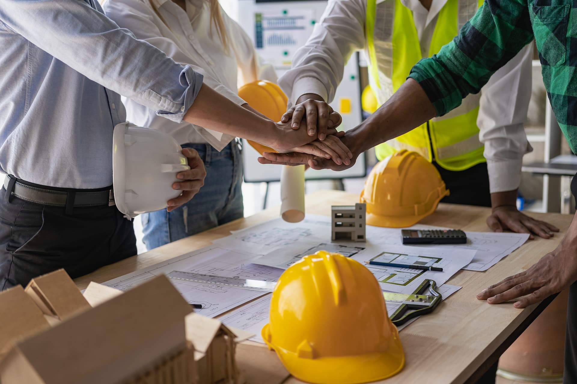 Contractors in office having team huddle with hands in the middle over blueprints on table.